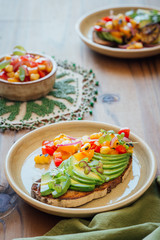 Top shot rye avocado toast, table setting for two, sliced avocado, rustic salsa of tomato, yellow peppers, red onion and micro greens, side serving of salsa, natural wooden background 