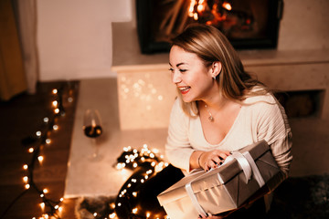 Portrait of an excited cute girl in casual clothes holding unopened present box by the fireplace