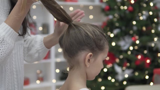Mom braids hair in the tail of her elegant daughter. Preparing for a great Christmas holiday. A girl in a lush white dress stands with her back to a woman, oa collects her hair with her hands in one t