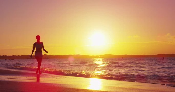 Sunset on beach with woman walking enjoying vacation in warm yellow sun wotj ocean waves and sea in slow motion.