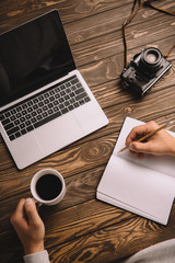 cropped view of male photographer writing in notebook at table with cup of coffee, laptop and retro...