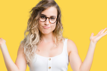 Beautiful young blonde woman wearing glasses over isolated background clueless and confused expression with arms and hands raised. Doubt concept.