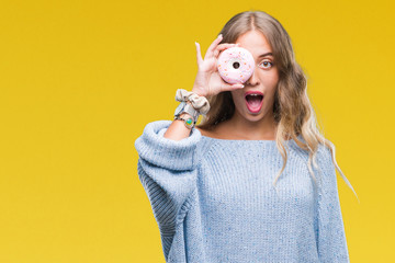 Beautiful young blonde woman eating pink donut over isolated background scared in shock with a surprise face, afraid and excited with fear expression