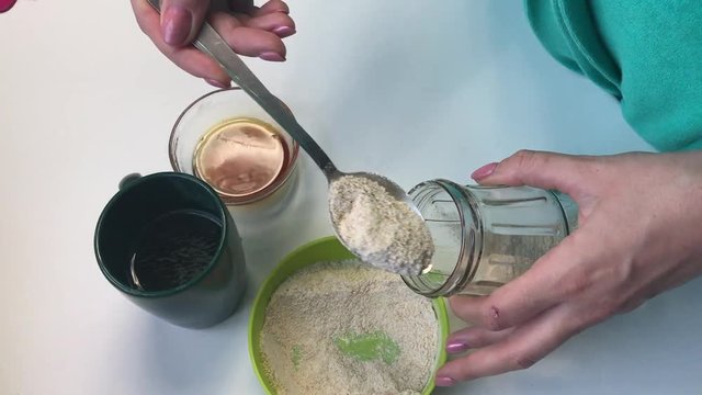 Woman puts a tablespoon of flour in a glass jar. For the preparation of yeast for baking bread at home. Other ingredients are located nearby on the table.