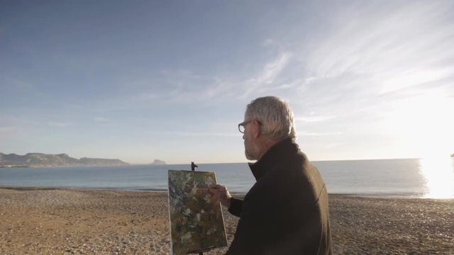 Senior man paints a picture on the beach. Elderly male artist makes the finishing touches on the modern abstract painting against raising sun over seashore. Morning sun reflected in the calm sea water