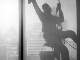 Silhouette images and shoot through transperent curtain effect from inside building which a man cleaning the window of high office building with his equipment such as wipe, sponge, bucket 