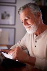 Senior Hispanic man sitting at a table using a tablet computer in the evening, close up, vertical