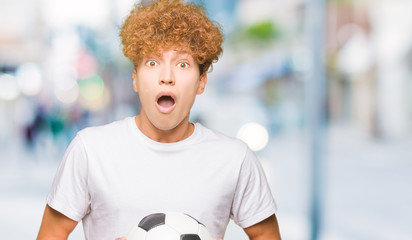 Young handsome man holding soccer football ball scared in shock with a surprise face, afraid and...