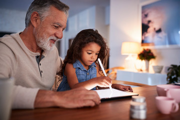 Senior Hispanic man at home with his young granddaughter using stylus and tablet computer