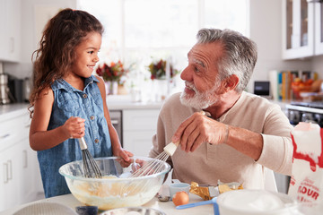 Young girl and grandfather making cake mixture at the kitchen table, smiling at each other, close up