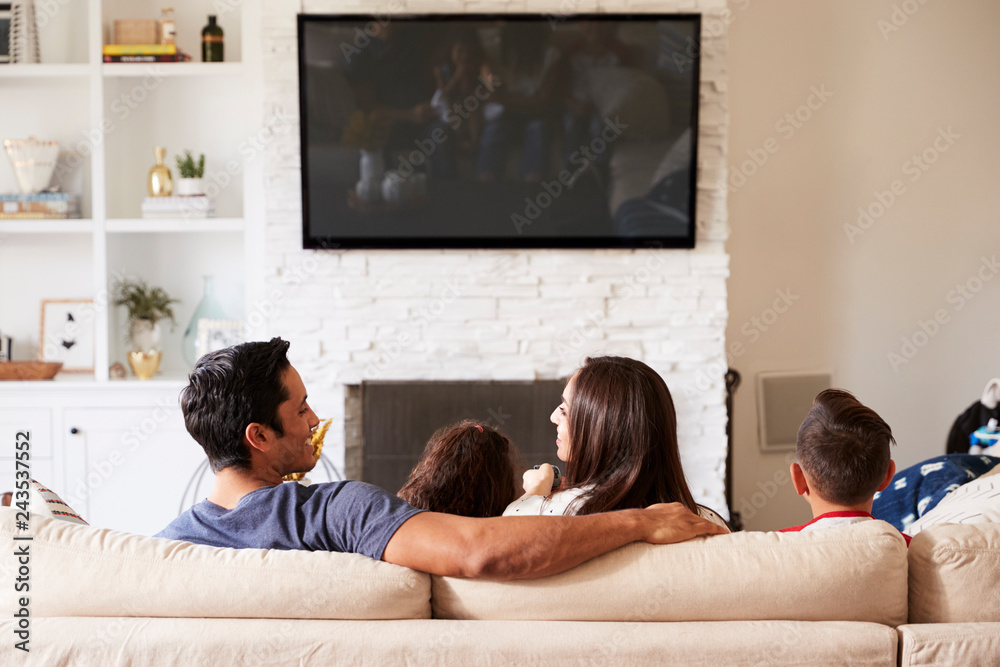 Wall mural Back view of young Hispanic family of four sitting on the sofa watching TV, mum looking at dad