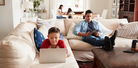 Pre-teen boy lying on sofa using laptop, dad sitting with tablet, mum and sister in the background