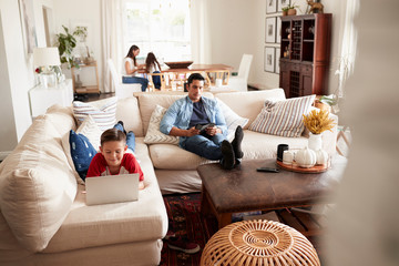 Pre-teen boy lying on sofa using laptop, dad sitting with a tablet, mum and sister in the background