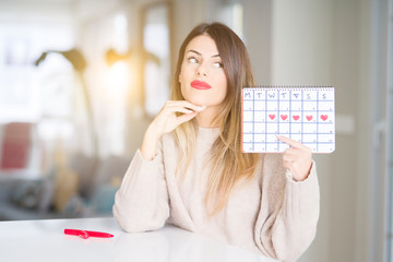 Young beautiful woman holding menstruation calendar at home serious face thinking about question,...