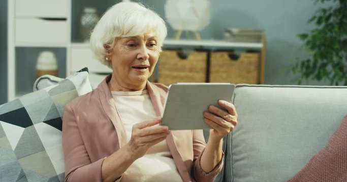 Caucasian senior grey haired woman having a videochat on the tablet device via webcam in the nice living room. Indoor.