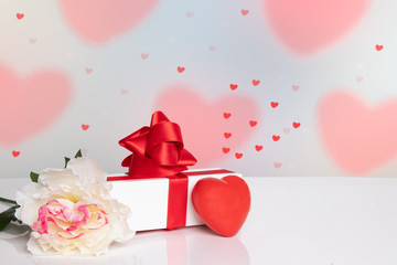 Valentine Day background. A bright table top with a flower, gift box and red heart in front of abstract lovely red hearts background. Template for your product display montage. Space.