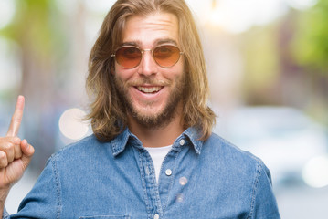 Young handsome man with long hair wearing sunglasses over isolated background pointing finger up with successful idea. Exited and happy. Number one.