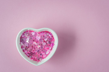 Fototapeta premium Sparkles in heart shaped bowl, ceramic cup on pink background. Concept about love and relationship. Valentines Day background with hearts. Copy space, flat lay.