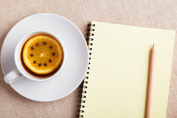 A Cup of tea with a piece of orange and cloves next to a pencil and a notebook on a table covered with a linen tablecloth, write down the list of actions. Concept of planning. Top view, copy space