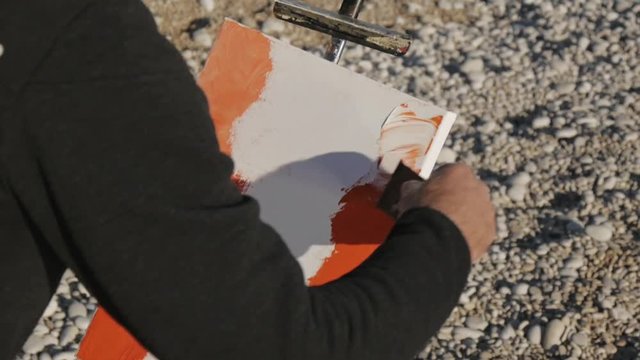 Senior man paints a picture on the beach. Elderly male artist applying acrylic paint to canvas with wide spatula at pebble sunrise sea beach.