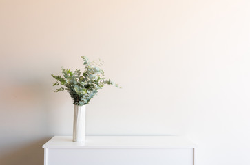 Eucalyptus leaves in tall white vase on sideboard against neutral wall background with copy space (selective focus)