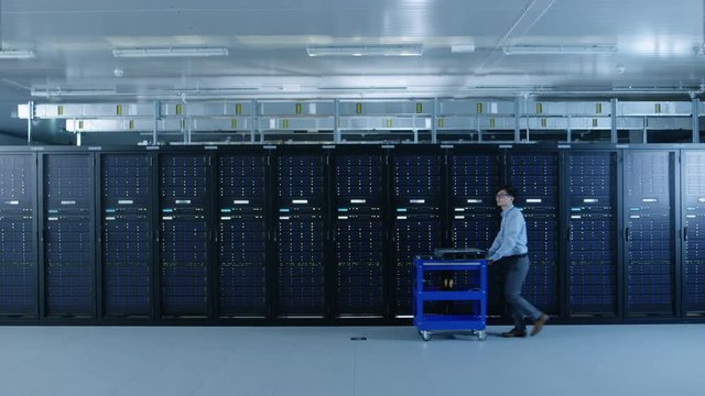 In the Modern Data Center: IT Technician Working with Server Racks, Pushes Cart Between Rows of Server Racks. On a Pushcart New Hardware for System Update. Engineer Doing Maintenance and Diagnostics.