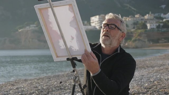 Senior man paints a picture on the beach. Elderly male artist in glass applying paint on white canvas on metal easel at sunset sea beach.