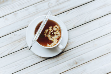 A mercury glass thermometer lies on a white cup with a linden, fragrant healing tea made from dried linden flowers on a grey wooden background. The concept of herbal medicine.