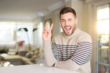 Young handsome man at home smiling with happy face winking at the camera doing victory sign. Number two.