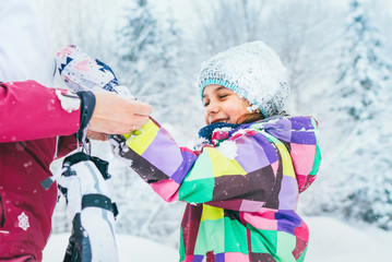 Mother helping her little daughter wearing warm gloves during snowy forest walking.