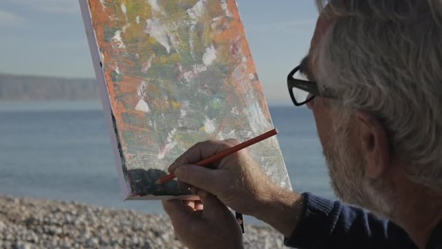 Senior man paints a picture on the beach. Elderly male artist signing his modern abstract picture on morning calm sea background. Close-up.