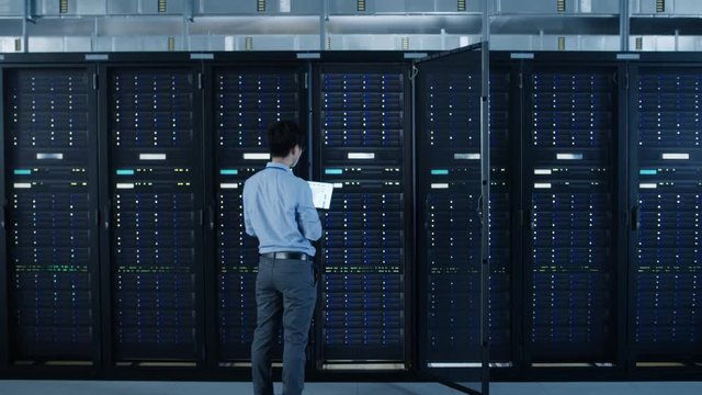  In the Modern Data Center: IT Engineer Standing Beside Open Server Rack Cabinets, Does Wireless Maintenance and Diagnostics Procedure with a Laptop. Zoom Out Camera Shot.