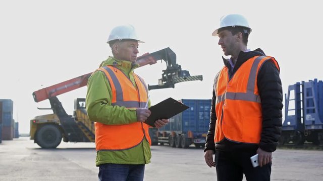 Two engineers wearing hardhats talking together while standing on a large commercial shipping dock tracking inventory