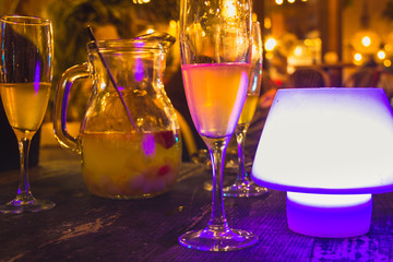 Cups and champagne sangria pitcher on wooden table by purple light lamp at night club. Celebration at restaurant bar, party atmosphere concepts