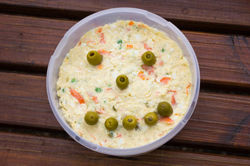 Delicious potato salad plastic bowl with smiling face made out of green olives on wood planks table. Fresh vegetarian meal concept