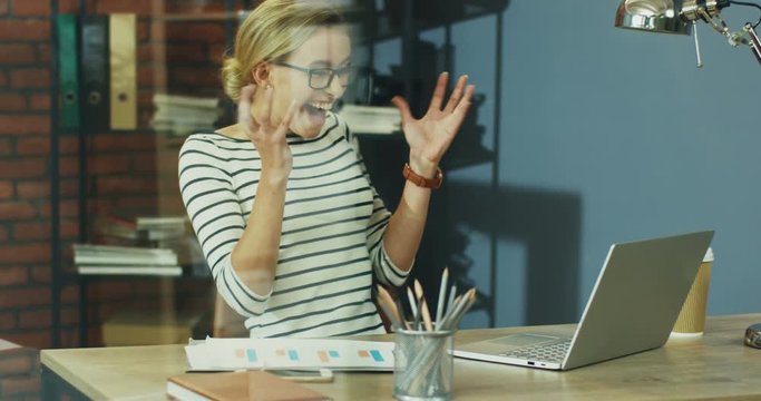 Young pretty Caucasian blond woman in glasses working at the computer in the office and then being very cheerful and happy as seeing some great news or work result.