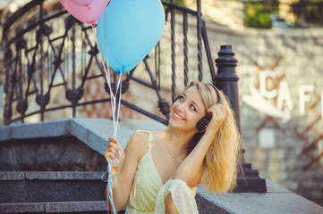 Beauty girl with colorful balloons laughing on city steps. Beautiful happy young woman on a sunny bright day. Joyful model is listening to music and celebrating with a pastel color balloon