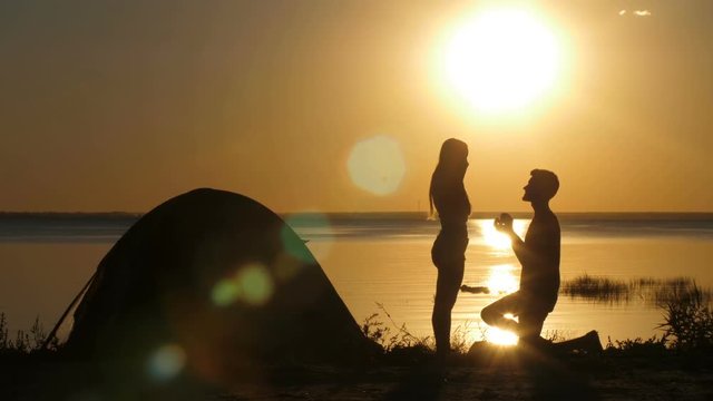 Silhouettes of kneeled man proposing his beloved woman on the beach at sunset. Boyfriend putting engagement ring on happy surprised female's finger. Couple embracing in happiness