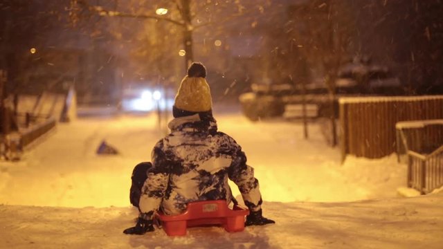 Cute young boy sledging in the evening on a snowy winter night, street lights on