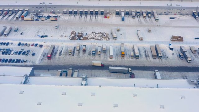Aerial look-down shot of a logistics loading dock with a semi-trailers trucks standing at warehouse ramp on load/unload goods and a lot of commercial carrier cars standing on the parking lot 