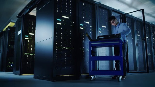 In Data Center: Male IT Technician Running Maintenance Programme on a Laptop, Controls Operational Server Rack Optimal Functioning. Modern High-Tech Telecommunications Operational Super Computer.