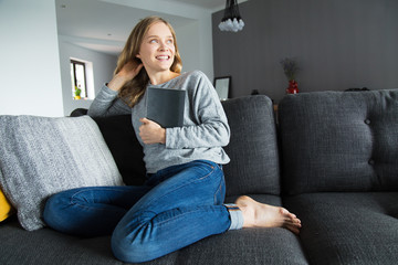 Cheerful female student done with her home assignment. Young woman in casual sitting on couch in living room and holding notebook. Happy bookworm concept