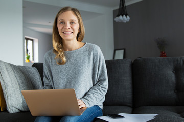 Positive freelance worker posing in her living room. Beautiful young woman on couch holding laptop...