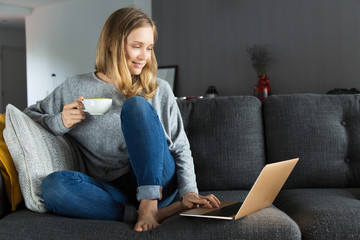 Positive girl enjoying weekend at home. Peaceful young woman sitting on couch in living room, holding white cup and smiling at laptop monitor. Residential Internet concept
