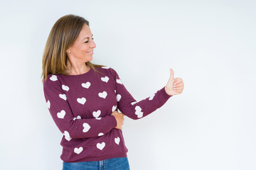 Beautiful middle age woman wearing heart sweater over isolated background Looking proud, smiling doing thumbs up gesture to the side