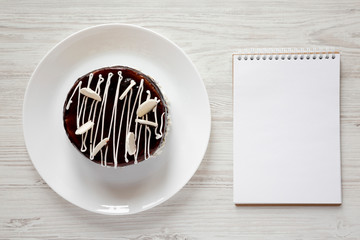 Homemade chocolate cherry cake and blank notepad on white wooden background, view from above. Flat lay, top view. Space for text.