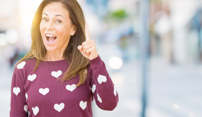 Beautiful middle age woman wearing hearts sweater over isolated background pointing finger up with successful idea. Exited and happy. Number one.
