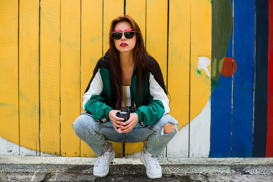 Portrait of a girl in clothes in the style of the 90s, sporty style, jacket, jeans, bananas, sunglasses. Lady on the background of wooden colored background, drinking morning coffee