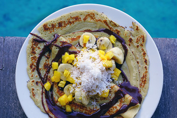 A tropical breakfast crepe with pineapple, chocolate, and fresh coconut eaten by the blue sea