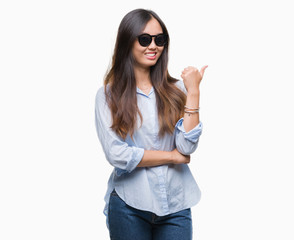 Young asian woman wearing sunglasses over isolated background smiling with happy face looking and pointing to the side with thumb up.